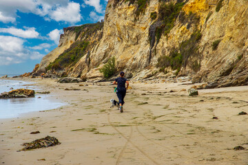 a woman running along the silky brown sands of the beach surrounded brown cliffs covered in lush green trees and plants with ocean water and blue sky with clouds at Leadbetter Beach in Santa Barbara - obrazy, fototapety, plakaty