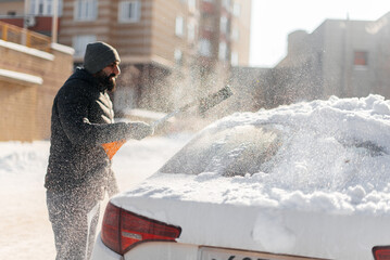A young man cleans his car after a snowfall on a sunny, frosty day. Cleaning and clearing the car from snow on a winter day. Snowfall, and a severe snowstorm in winter.