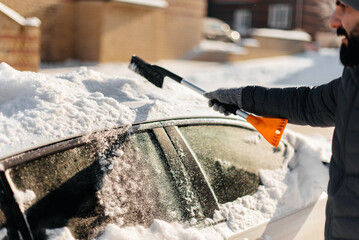 A young man in close-up cleans his car after a snowfall on a sunny, frosty day. Cleaning and clearing the car from snow on a winter day. Snowfall, and a severe snowstorm in winter.