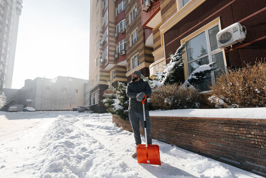 A man stands with a shovel and removes snow in front of the house on a sunny and frosty day. Cleaning the street from snow on a winter day. Snowfall, and a severe snowstorm in winter.