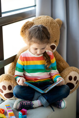 A cute girl of 6 years old sits on the couch by the window with a large toy bear and reads a book....