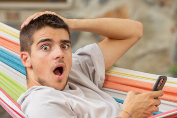 surprised and amazed young man with mobile phone in hammock