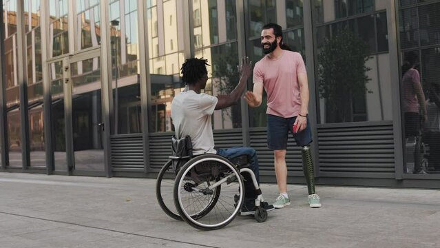 Multiracial friends with physical disability hugging each other outdoor - Man with leg prosthesis greeting friend sitting in wheelchair