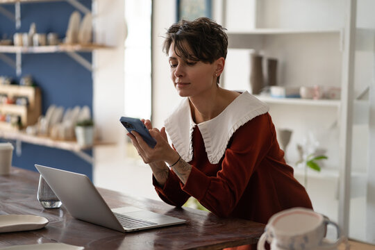 Young woman pottery store manager using mobile phone at workplace, female ceramics artist connecting with customer through social media, craftswoman with smartphone promoting craft business online