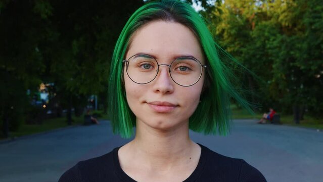 Portrait of nice girl with green short hair in glasses. Young woman looking at camera, smiling in city park. Gaze of lady in black with grey blue eyes. Front view