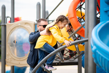 A girl 6-7 years old with her dad on the playground. She on the slide and has a fun. Dad helps the girl climb the stairs. Childhood. Time with family. Spring. City.
