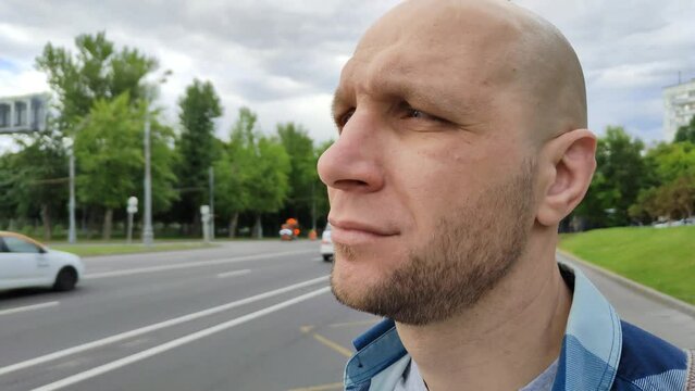 Young man standing near highway, waiting for bus. Cute bald guy at public transport stop. Man in plaid shirt wait for taxi, standing at roadside, looks at road along which cars and trucks traveling