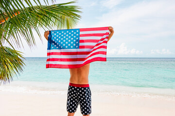 young athletic man stands in swimming trunks with an American flag on his shoulders near ocean sea shore, concept of celebrating America's USA Independence Day on July 4 and patriotic education, sense