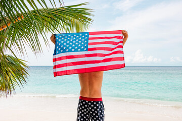 young athletic man stands in swimming trunks with an American flag on his shoulders near ocean sea shore, concept of celebrating America's USA Independence Day on July 4 and patriotic education, sense