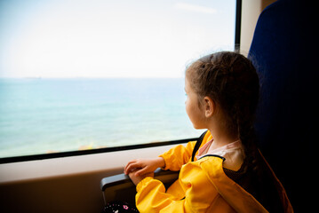 A little girl of 6-7 years old looks out the window of a train at the sea. She's wearing a yellow parka. Journey. Reflection. Vacation. Summer. Family vacation.