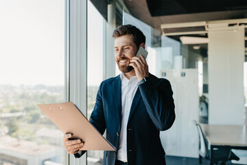 Happy businessman talking on smartphone and checking papers, standing in modern office interior, free space