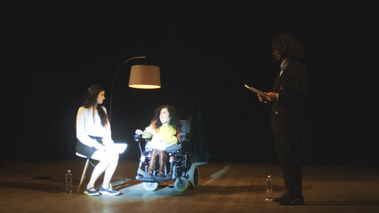 Theater actors with a disabled actress in a wheelchair emotionally playing roles on a stage illuminated by a spotlight during a play rehearsal