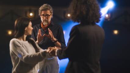 An actress and an actor rehearsing a love scene on a theater stage with a spotlight while a male director watching them