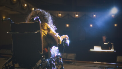 A disabled actress with spinal muscular atrophy in a wheelchair on a stage illuminated by a spotlight talking with the director in the theater