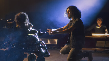 An actor singing to actress with a disability in a wheelchair while playing a romantic scene on the stage of the theater with a spotlight, during a rehearsal with a director.