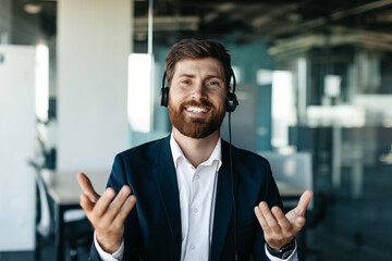 Portrait of happy businessman in headset talking and gesturing at camera, sitting in office interior, copy space