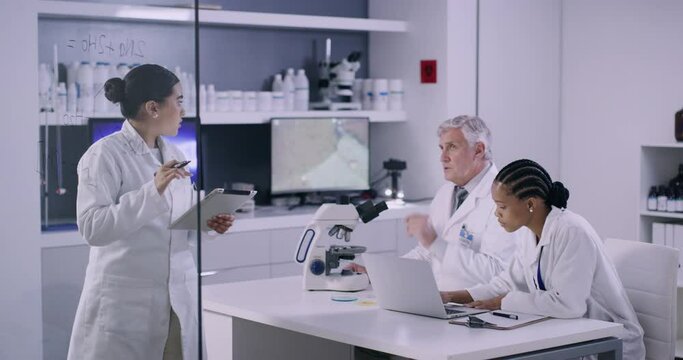 Team of scientists in a medical research lab discussing a chemical formula for a vaccine. Female researcher writing on a glass wall with colleagues working at a table in a modern laboratory
