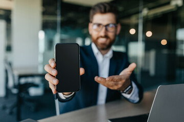 Happy businessman showing smartphone with blank screen, sitting at modern office, mockup for mobile...