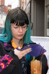 natural real person photo, a young woman wearing glasses  looking at her mobile phone whilst drinking a cocktail on a busy shopping street. Urban lifestyle concept