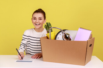 Happy woman office worker sitting at workplace with cardboard box with her things and smiling,...