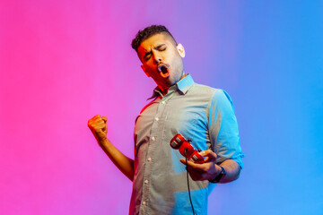 Extremely satisfied man in shirt playing video game, holding joystick, celebrating his winning, clenched fist and yelling happily. Indoor studio shot isolated on colorful neon light background.