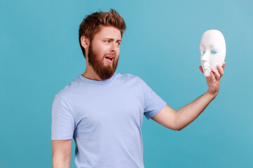 Portrait of confused puzzled handsome young adult bearded man holding white mask in hands and looking with open mouth, despises hidden personality. Indoor studio shot isolated on blue background.