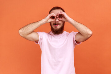 Fototapeta na wymiar Portrait of bearded man with funny face looking through fingers shaped like binoculars and expressing positive, wearing pink T-shirt. Indoor studio shot isolated on orange background.