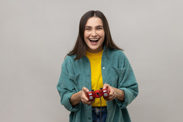 Excited woman holding gamepad, winning video games competition, looking at camera with happy expression, wearing casual style jacket. Indoor studio shot isolated on gray background. © khosrork