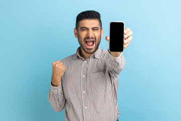 Obraz na płótnie Canvas Happy businessman with beard holding smartphone and smiling making yes gesture, celebrating online lottery or giveaway victory, wearing striped shirt. Indoor studio shot isolated on blue background.
