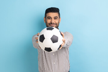 Happy handsome bearded businessman holding out soccer ball on his hand with smiling positive expression, wearing striped shirt. Indoor studio shot isolated on blue background.