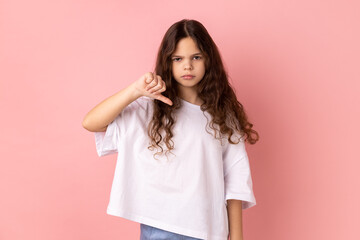 Portrait of little girl in white T-shirt criticizing bad quality with thumbs down displeased grimace, showing dislike gesture, expressing disapproval. Indoor studio shot isolated on pink background. - obrazy, fototapety, plakaty