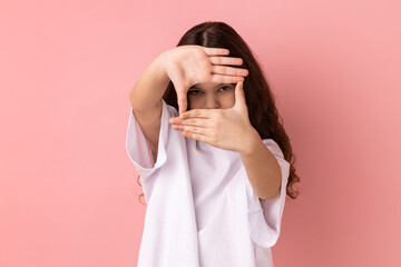 Portrait of concentrated little girl wearing white T-shirt looking at camera with one eye, focusing through photo frame made of fingers. Indoor studio shot isolated on pink background.