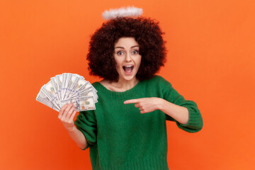 Amazed woman with Afro hairstyle wearing green casual style sweater and nimb over head holding big sum of money, pointing at dollar banknotes. Indoor studio shot isolated on orange background.