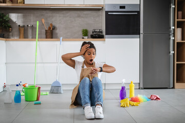 Shocked young black housewife with open mouth sit on floor with cleaning supplies, look at clock
