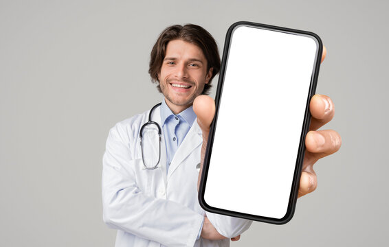Positive male doctor in medical coat showing mobile phone, mockup