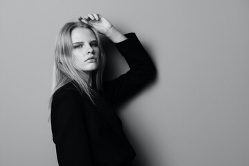 Serious young blonde woman leans on the studio wall holding hand overhead looks at camera posing isolated in black jacket