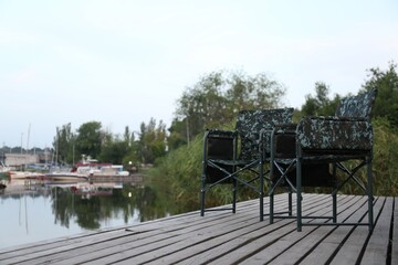 Camouflage fishing chairs on wooden pier near river, space for text