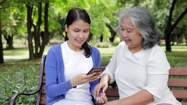 Elderly mother and Asian daughter lounging in the park Happy on vacation, holding smartphones and taking pictures together. family concept Health care for the elderly. senior caregiver