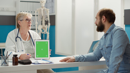 Obraz premium Doctor vertically holding greenscreen on digital tablet in cabinet. People at checkup visit looking at mockup template with isolated chromakey background and blank copyspace on display.