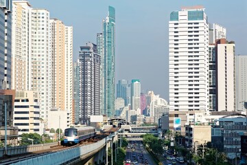 Obraz premium View of a BTS skytrain traveling on elevated metro system between high rise skyscrapers in Downtown Bangkok on a beautiful sunny day~ Cityscape of Bangkok, the fast developing capital city of Thailand