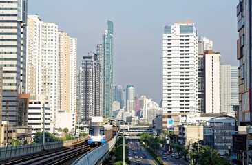 Naklejka na ściany i meble View of a BTS skytrain traveling on elevated metro system between high rise skyscrapers in Downtown Bangkok on a beautiful sunny day~ Cityscape of Bangkok, the fast developing capital city of Thailand