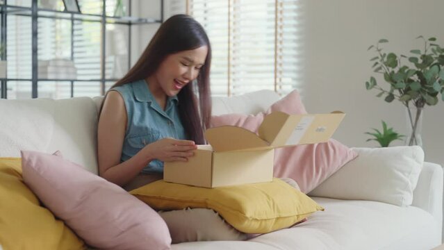 Young Gen Z influencer girl open gift postal mail box sit at home sofa couch. Asia people enjoy wow unbox carton parcel order online buy from small retail store SME shop supply chain omni channel.