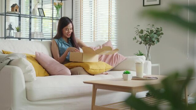 Young Gen Z influencer girl open gift postal mail box sit at home sofa couch. Asia people enjoy wow unbox carton parcel order online buy from small retail store SME shop supply chain omni channel.