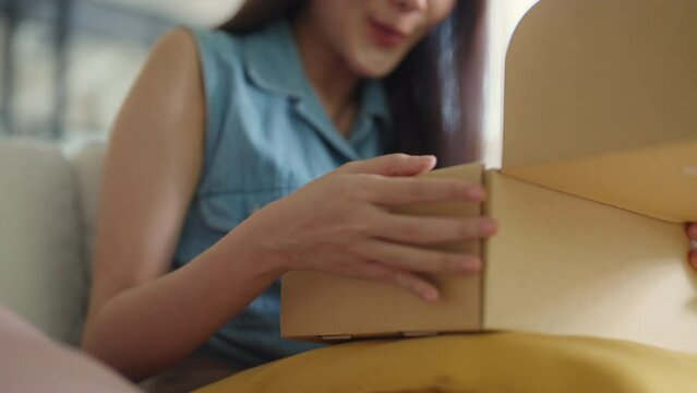 Young Gen Z influencer girl open gift postal mail box sit at home sofa couch. Asia people enjoy wow unbox carton parcel order online buy from small retail store SME shop supply chain omni channel.