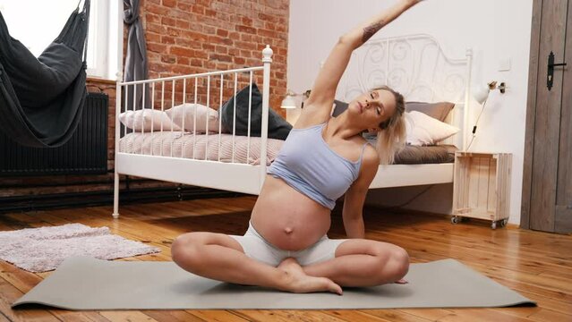 Beautiful caucasian pregnant woman in baby blue tank top sitting in cross-legged squat and stretching her side. Modern bedroom interior. High quality 4k footage