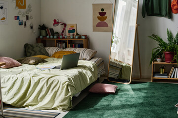 Horizontal image of modern teenage bedroom with bed and laptop on it with big mirror in centre of...