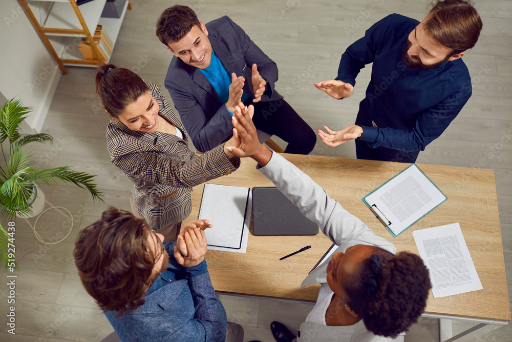 Wall mural Diverse team of people meet in office and make business deal. Two happy women stand up from table and give each other high five while colleagues are applauding. Top view. Partnership, success concept