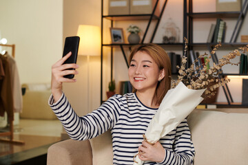 Fototapeta na wymiar Positive young Asian woman taking selfie with bouquet of dried flowers to post on social media