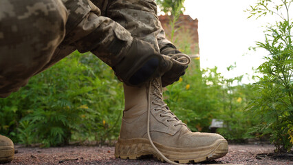 a soldier in a camouflage uniform and tactical gloves laces up his military boots. tactical...