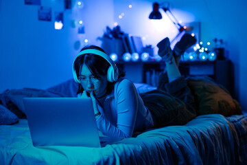 Teenage girl lying on her bed in wireless headphones and playing online on laptop in dark neon room © AnnaStills